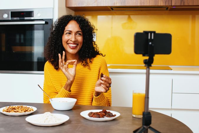 Woman in colorful kitchen live-streaming her breakfast of cereal, orange juice and dried fruit