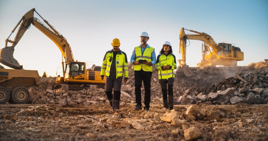 Construction Site With Excavators on Sunny Day: Diverse Team Of Male And Female Specialists Walking And Discussing Real Estate Project. Engineer, Architect, Urban Planner Talking, Using Tablet.