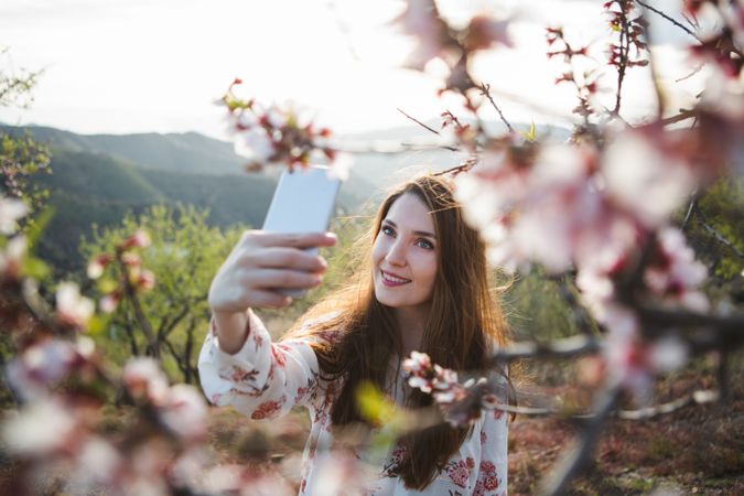 Young happy woman taking selfie on smartphone near trees on hill