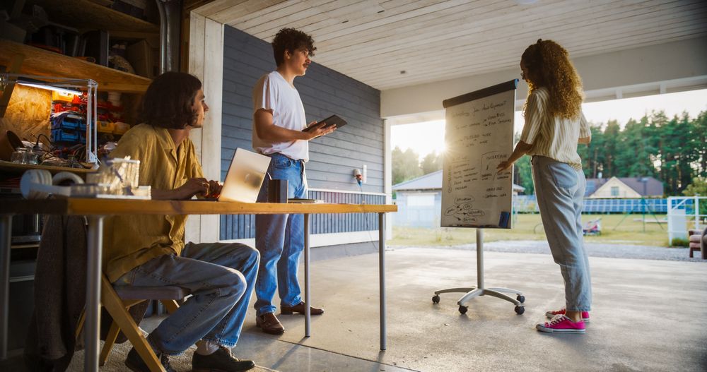 Hispanic Female CEO Using Whiteboard To Explain Business Objectives To Two White Male Colleagues With Laptop And Tablet.