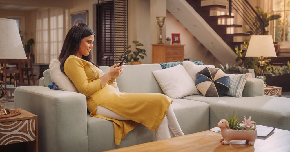 Young Indian Woman Browsing Smartphone On Sofa, Engaging In E-Commerce, Online Shopping
