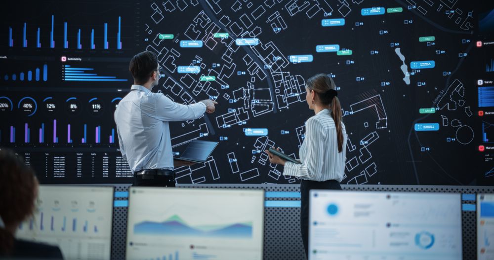 Male And Female Logistics Specialists Discussing New Routs Of Delivery In Front Of Big Digital Screen In Monitoring Office.