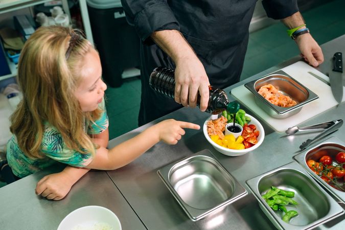 Chef pouring soy sauce on poke bowl while happy girl pointing at it in restaurant kitchen