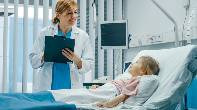 Recovering Little Girl Lies on a Bed In the Hospital, Friendly Doctor With Clipboard Asks Questions
