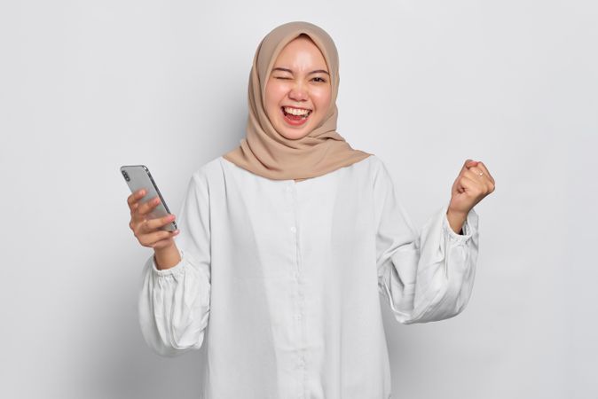 Excited Asian female in headscarf happy with arms up while holding her cell phone