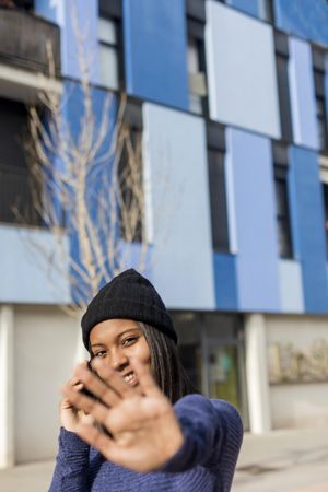 Female in wool hat and blue sweater blocking camera with hand while chatting on cell phone