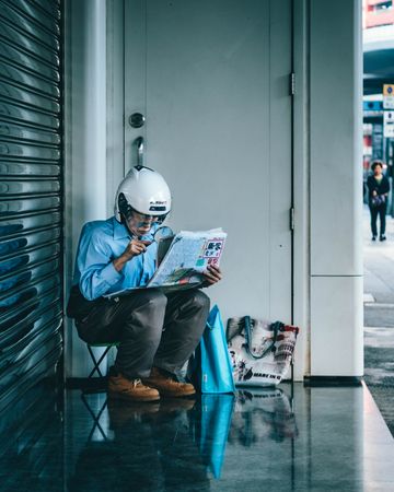 Older man with helmet sitting on a sidewalk reading newspaper