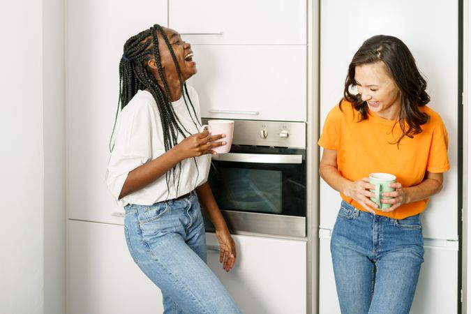 Two women laughing with each other while leaning on the fridge with coffee