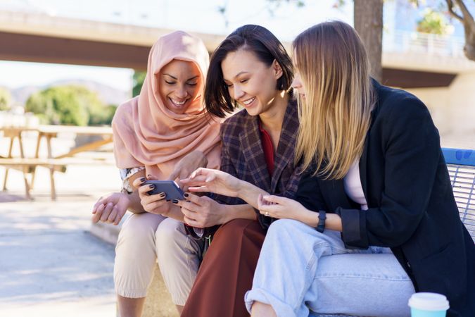 Three smiling women sitting on outdoor park bench talking while watching smartphone