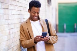 Laughing Cuban male leaning against wall texting on his phone