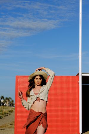 A long hair fashion model with a hat at the beach in front of a red wall