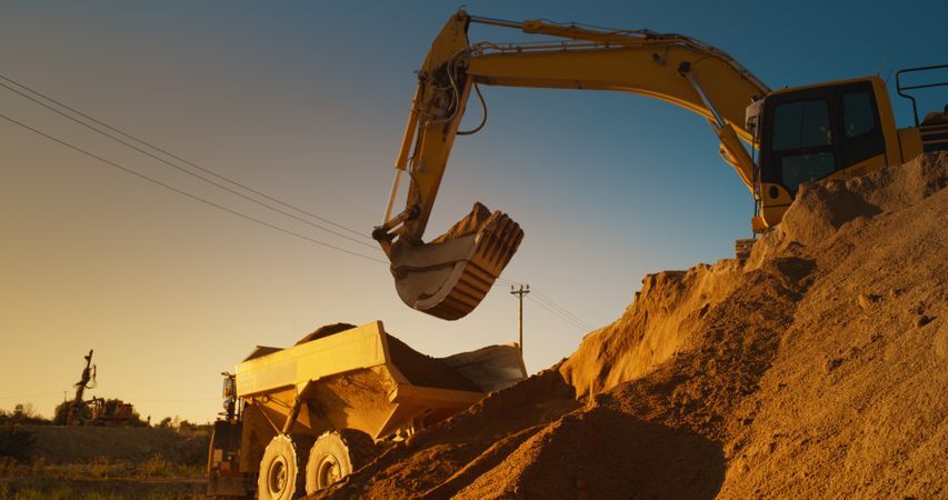 Construction Site On Sunny Evening: Industrial Excavator Loading Sand Into A Truck. The Process Of Building New Apartment Block. Workers Operating Heavy Machinery To Complete New Real Estate Project.