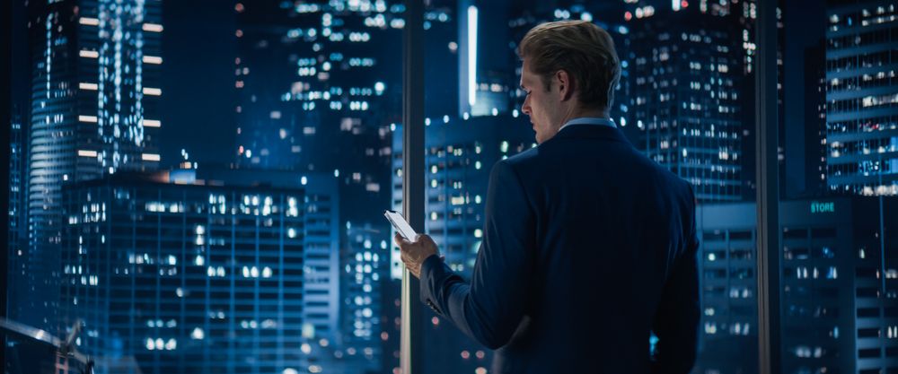 Backview of Businessman in Stylish Suit Standing Next to Window in Big City Office with Skyscrapers Late At Night.