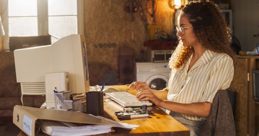 Side View Of Hispanic Female Engineering Student Writing Academic Thesis On Old Desktop Computer In Retro Garage.