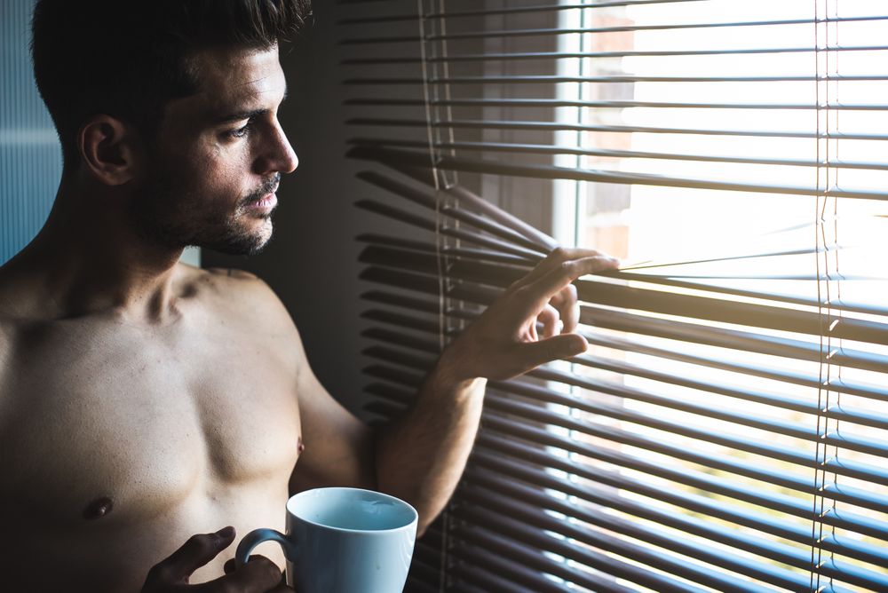 Man standing by a windows blind with a cup of coffee