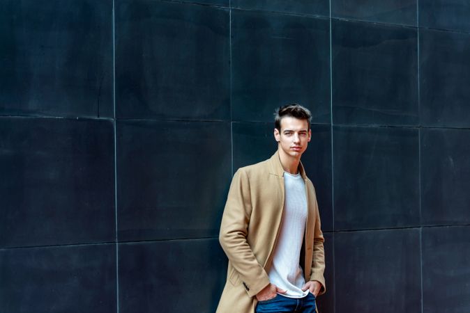 Young man leaning on dark wall outside looking at camera dressed in camel coat