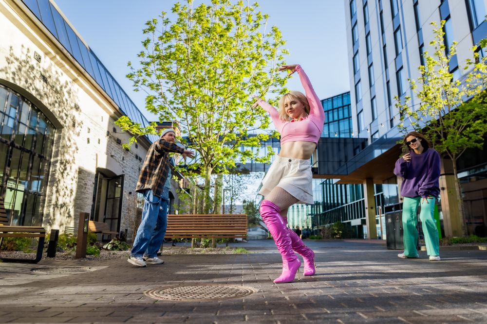 Vibrant Outdoor Scene Of Young Adults Dancing In An Urban Setting, With A Woman In Pink Leading Energetically.