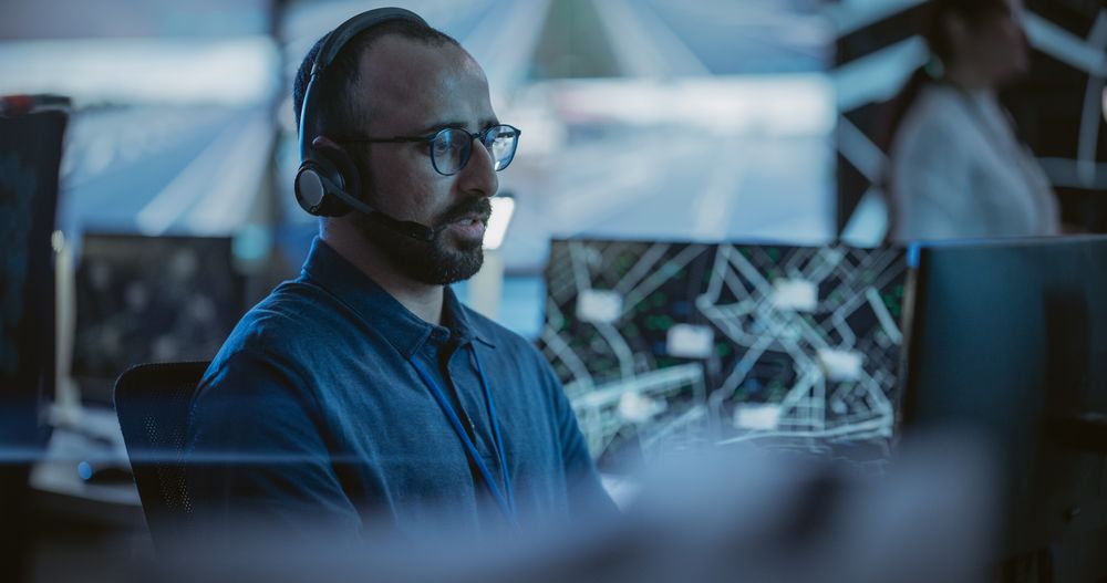 Multiethnic Male Officer Works on a Computer with Surveillance CCTV Video Footage in a Traffic Monitoring Center with Multiple Feeds on a Big Digital Screen.