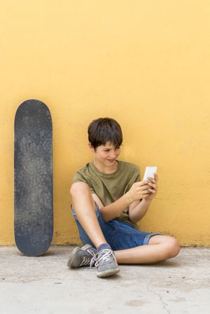 Teenage boy sitting on ground leaning on a yellow wall and taking a selfie