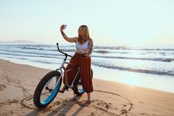 Cheerful woman taking selfie on smartphone while resting with bicycle at sunny shore