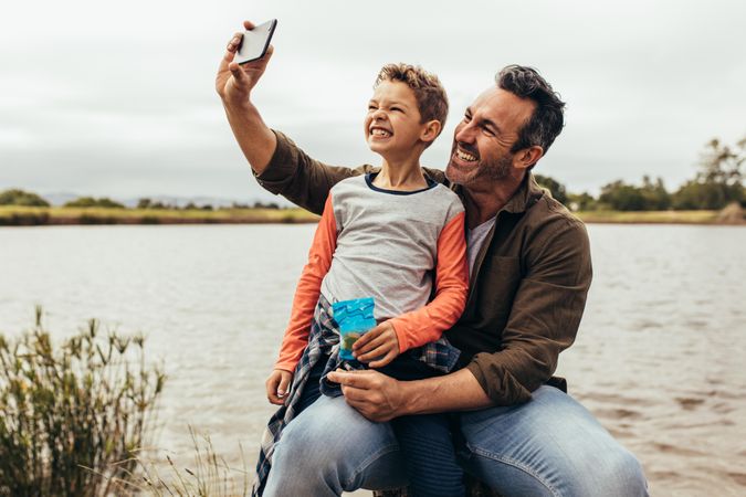 Happy man taking a selfie with his son sitting near a lake