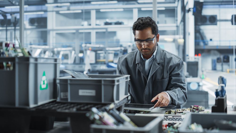 Indian Male Worker Disassembling Old Laptops To Recycle Electronic Components For Microchip Production At an Automated Electronics Factory With Robotic Arms.