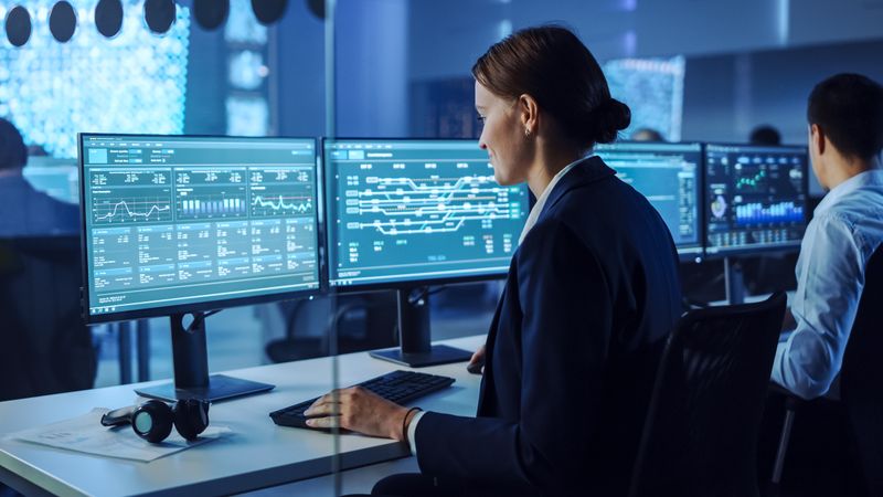 Data Analyst Examines Financial Charts on Dual Monitors in Dimly Lit Control Room.