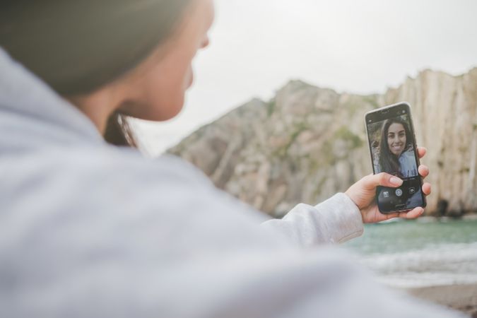 Woman taking selfie on smartphone on shore near stones and water