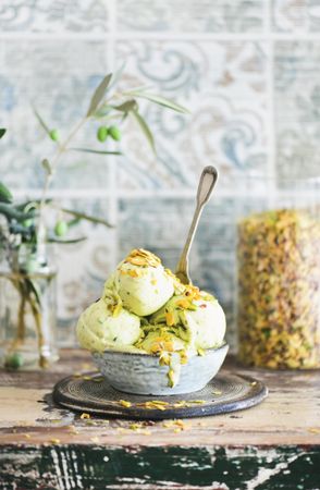 Bowl of melting pistachio ice cream with spoon and tiled background with leaves and glass container
