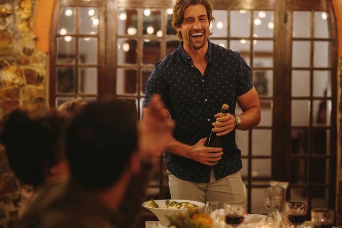 Laughing man opening champagne with friends sitting around dinner table