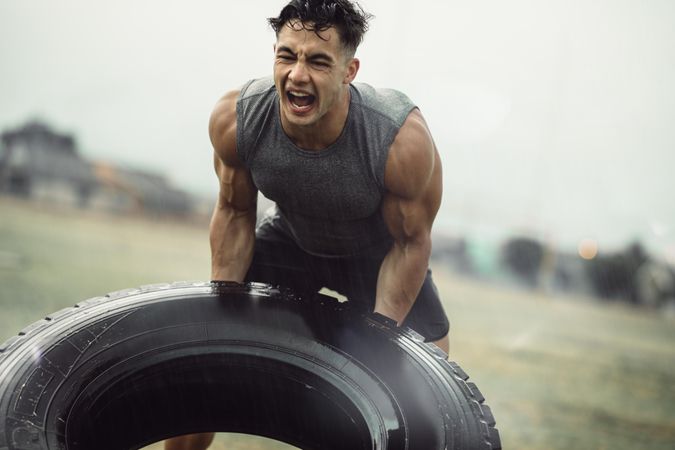 Tough young male athlete doing a tire flip exercise in the rain