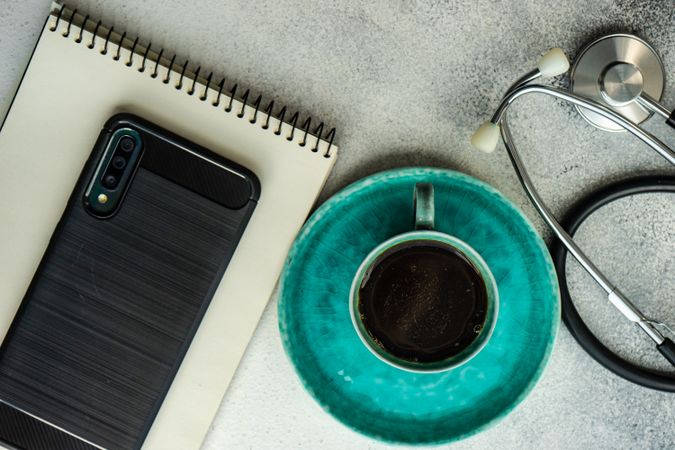 Top view of coffee cup on teal plate, phone, stethoscope and notepad