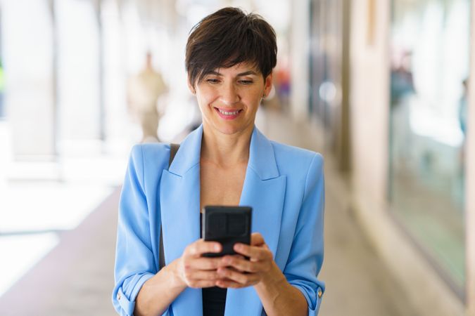 Happy professional woman in blue blazer checking phone while walking down street