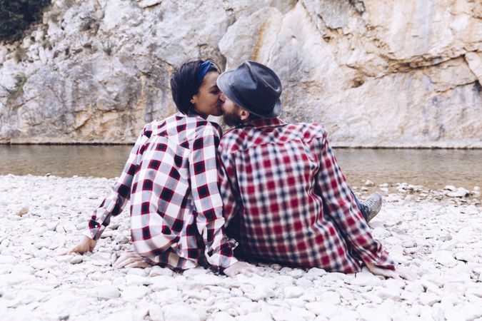 Positive woman and man resting and kissing on stone shore near river
