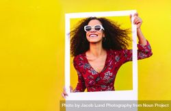 Woman with curly hair wearing sundress and sunglasses looking through blank photo frame bYVWX0
