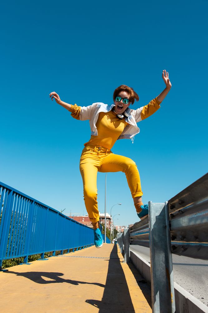 Stylish young carefree woman in vibrant clothes having fun on street bridge against blue sky