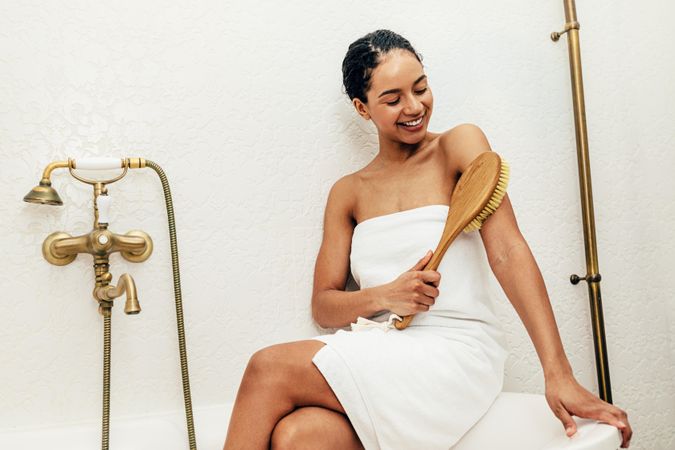 Woman wearing a towel sitting on edge of bath exfoliating with a brush