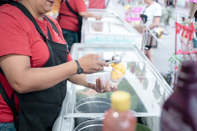 An Hispanic adult woman is serving some vanilla ice cream balls in a cup in the street