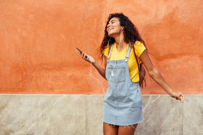 Dancing Arab woman with curly hairstyle using phone and headphones in front of on orange wall
