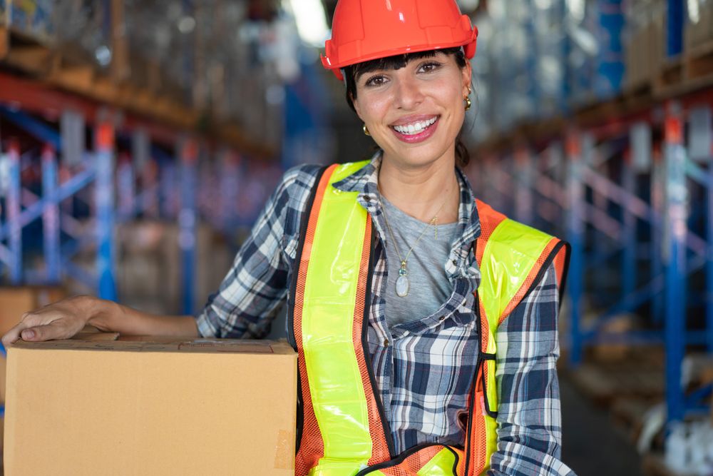 Woman in hard hat smiling and carrying in box in shipping center