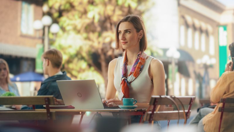 Female Using Laptop Computer on a Terrace in a Cozy Cafe on a Summer Day. Young Woman Working Remotely while Diverse People Sitting Behind Tables, Enjoying a Weekend in the City