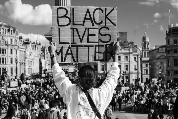 Grayscale back view of woman holding a banner that says " Black Lives Matter" against a crowd in London