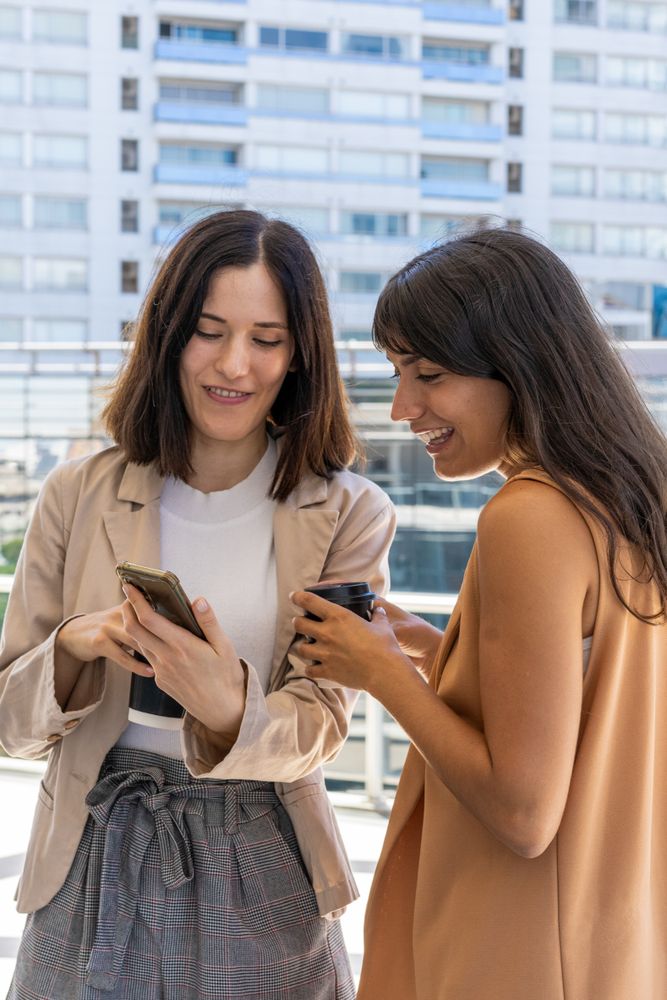 Two women are outside. One shows something on her phone while the other holds a cup
