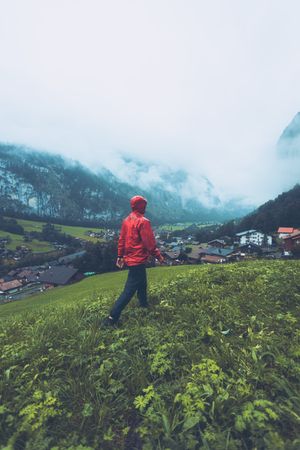 Back view of man in red jacket walking on green grass field near village in Switzerland