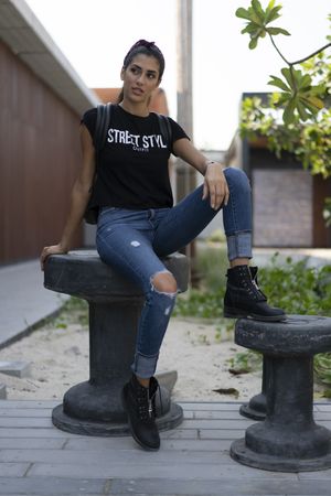 Young woman in denim pants and dark top sitting on concrete table outdoor