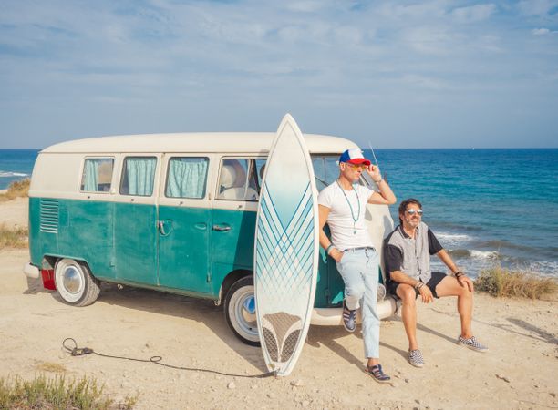 Men with surfboard near car on dusty seaside