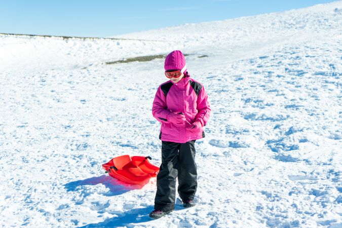 Child in pink snow suit tugging red sled behind her up a snowy hill