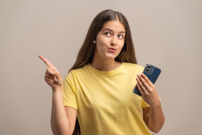 Thoughtful young woman holding a phone and pointing, considering an option.