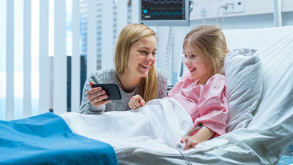 Cute Little Girl Lies on a Bed in the Children's Hospital, Her Mother Sits Beside, They Watch Cartoons/ Funny Videos on Smartphone