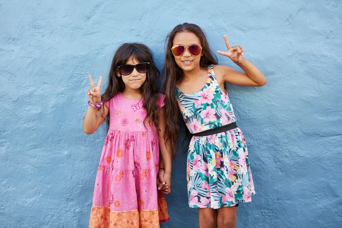 Portrait of two little girls standing together wearing sunglasses gesturing peace signs